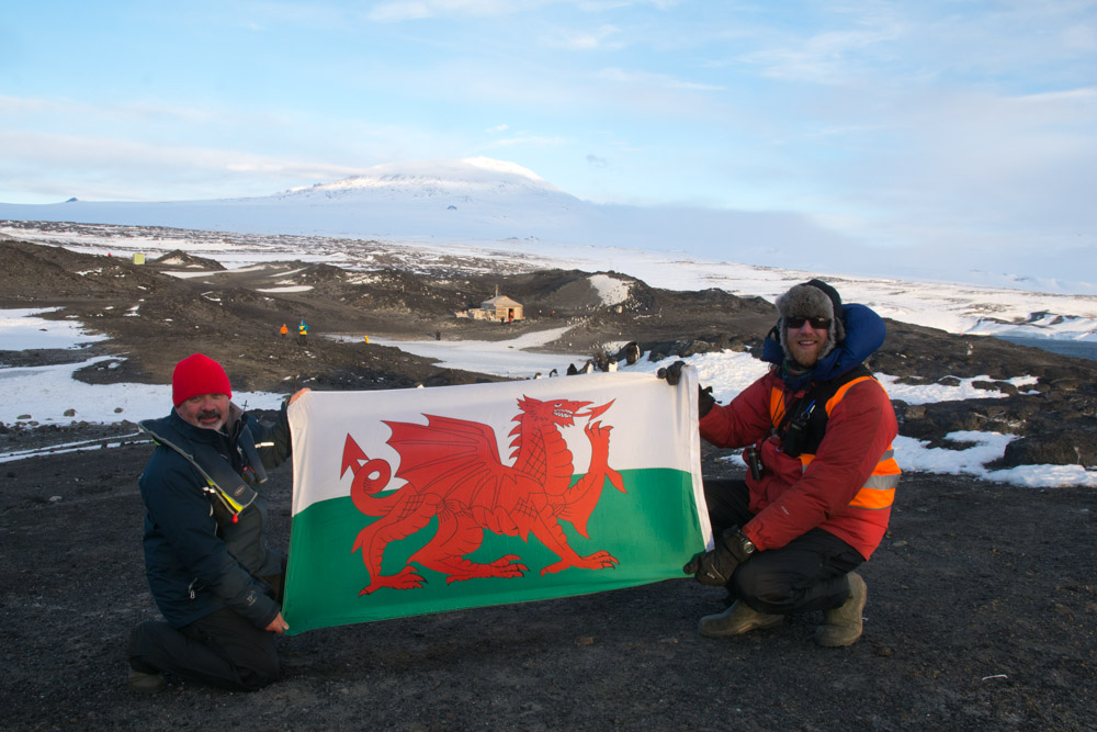 Myself, a friend and Welsh flag with Mount Erebus and Shackleton's hut, Cape Royds, Ross Island