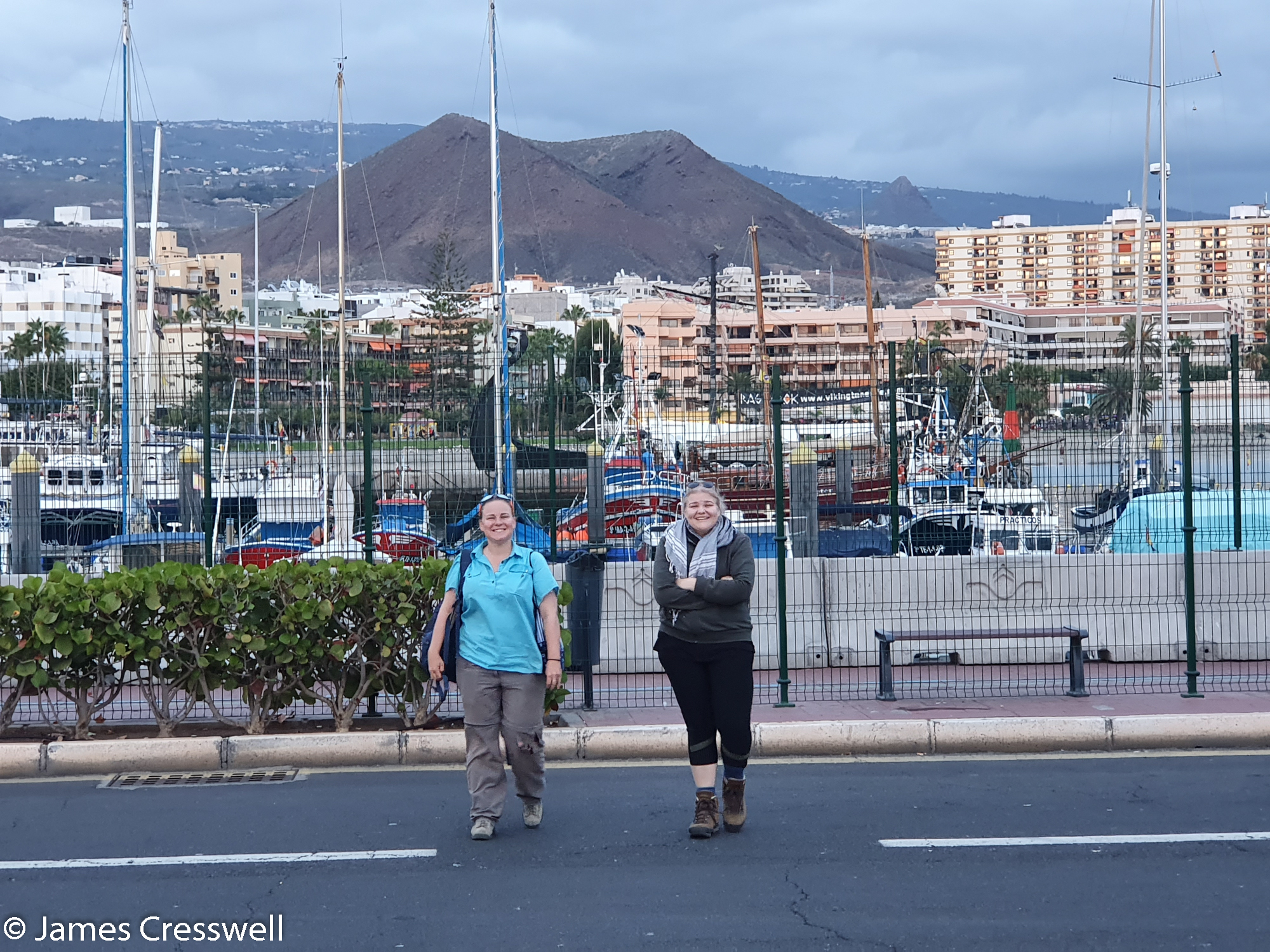 Two women crossing a road near a port with volanic feature behind
