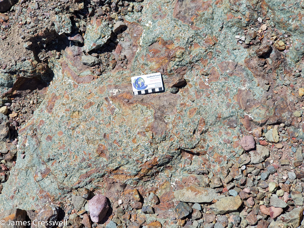 A photograph of a a green coloured rock that has red blotches and a scale card