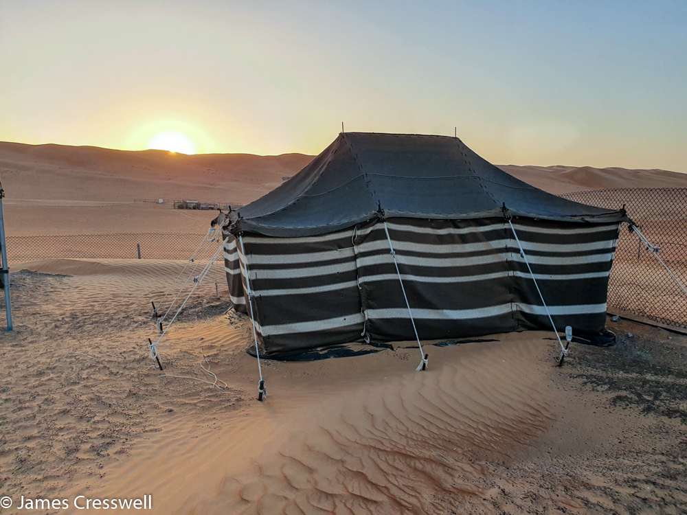 A photograph of a tent in the foreground, with the sun rising over sand dunes in the back ground