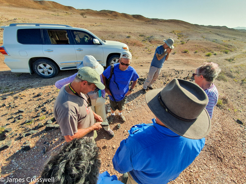 A photograph of a seven people talking about a rock, with a Landcruiser in the background