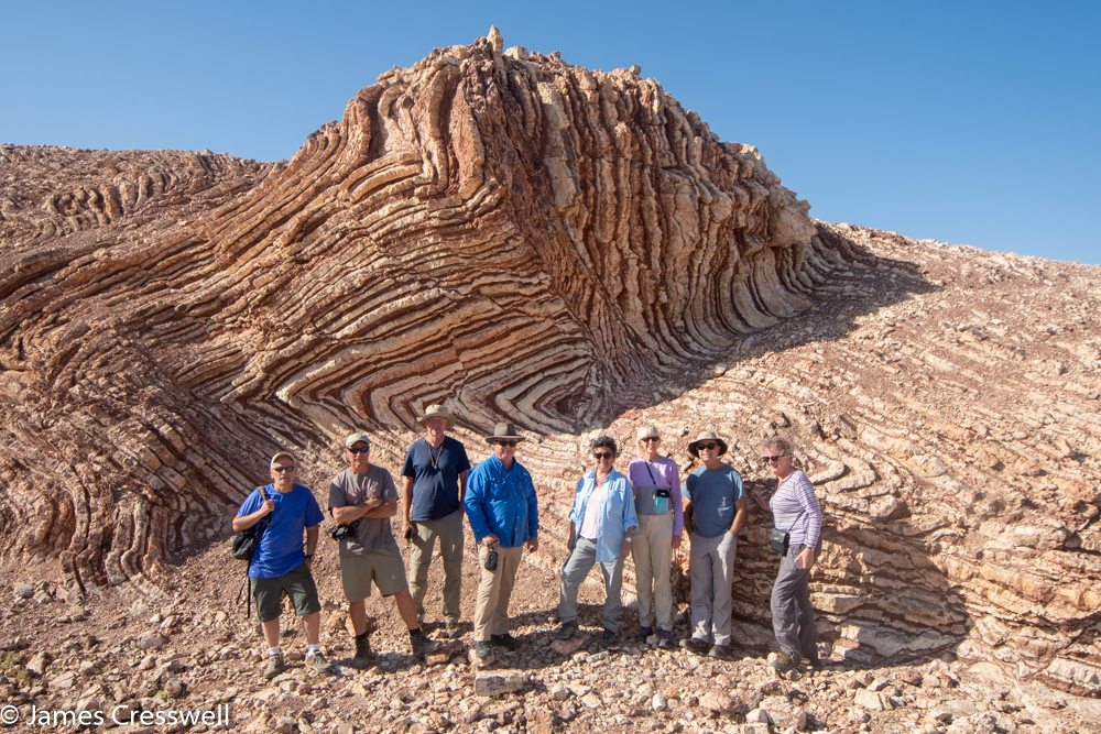 A photograph of a eight people standing in front of a small cliff that is made up of folded red and white layers