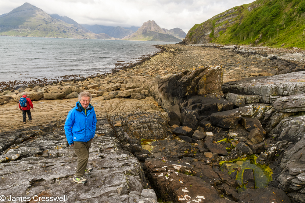 Two men stand on rocks with sea and mountains in the background