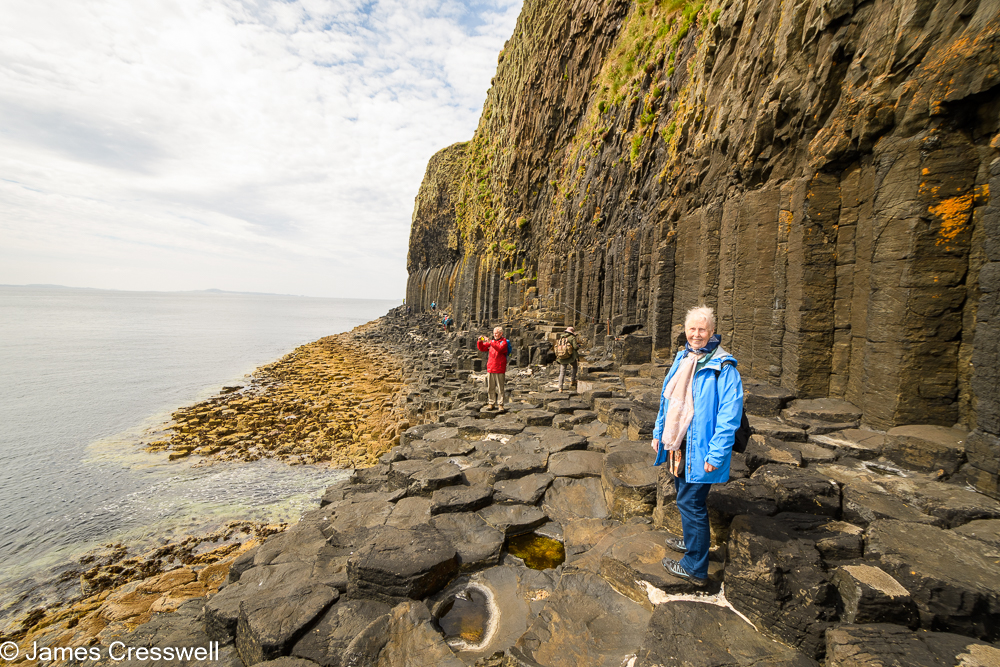 A woman stands on basalt columns, with a cliff of columnar basalt behind