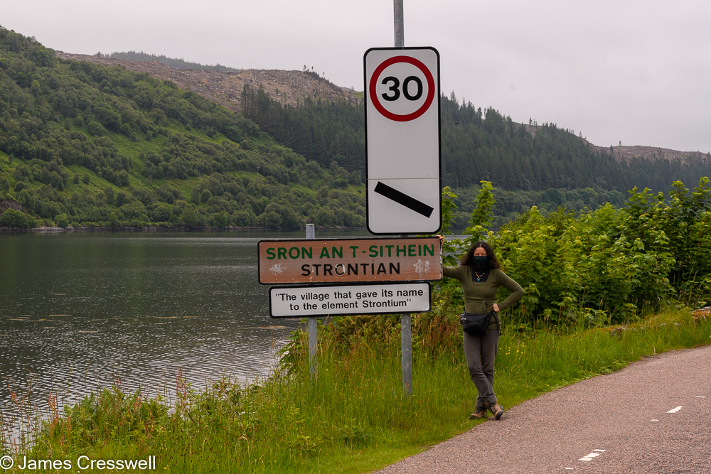 A woman standing next to a road sign that reads Strontian