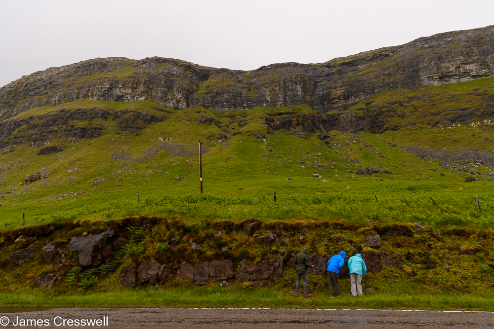 Three people examine rocks at the base of a cliff