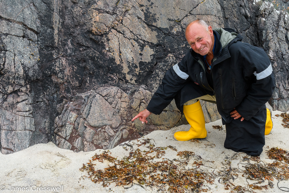 A man in yellow wellies points to brown colured veins in a grey and black rock