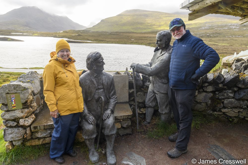 Two people with a statue of two men talking with a lake in the background